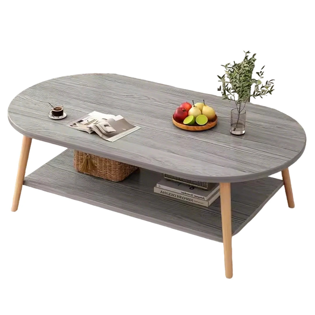 An oval coffee table with a gray wood-grain finish and light natural wood legs, featuring a lower shelf with books and a basket, styled with a fruit bowl, magazine, and vase on a white background.