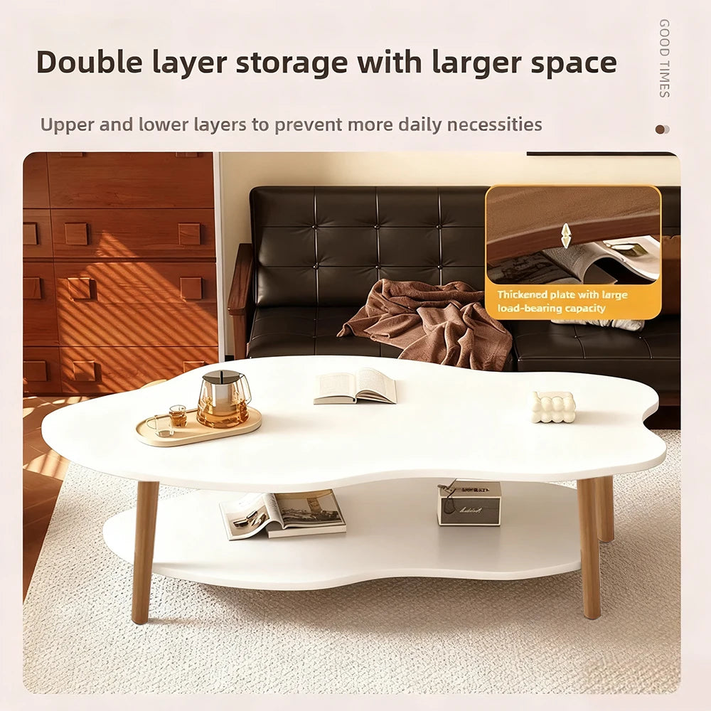 A modern white coffee table with a wavy organic silhouette, featuring a lower shelf and light wood legs, styled in a warm living room setting with a dark leather sofa and wooden cabinet in the background.