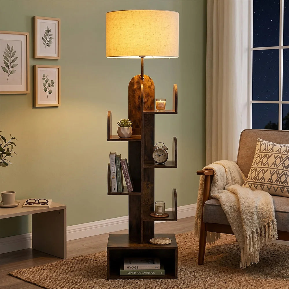 A tall, rustic wooden floor lamp styled in a cozy living room. The lamp features a warm beige fabric drum shade, a natural wood neck, and a dark brown tiered wooden stand with small decorative shelves holding books, a small plant, and candles.