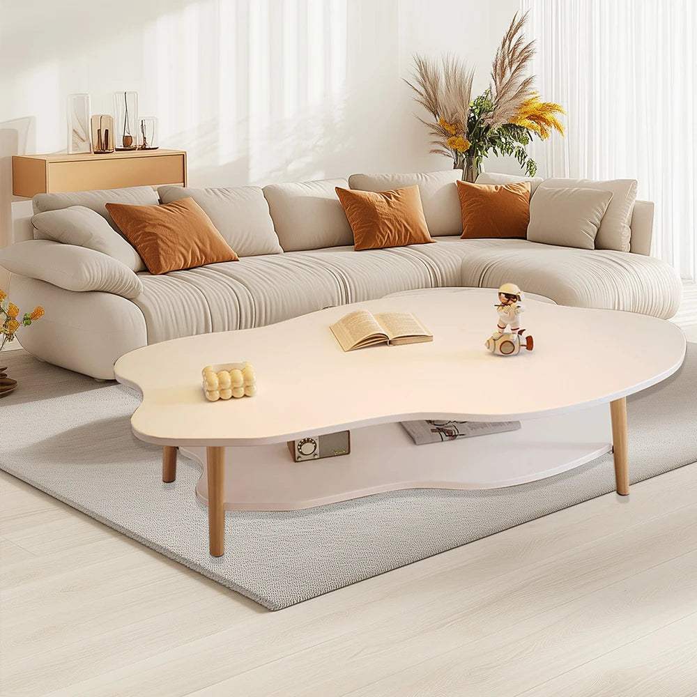 A modern living room scene featuring a cream-colored wavy coffee table with light wood legs, styled in front of a matching cream sectional sofa with orange accent pillows on a gray rug.