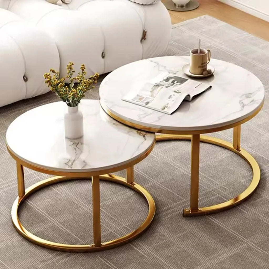 Two round nesting coffee tables with white marble-patterned tops and gold metal frames, styled in a living room setting with a white sofa, gray rug, vase with flowers, and a mug on a saucer.