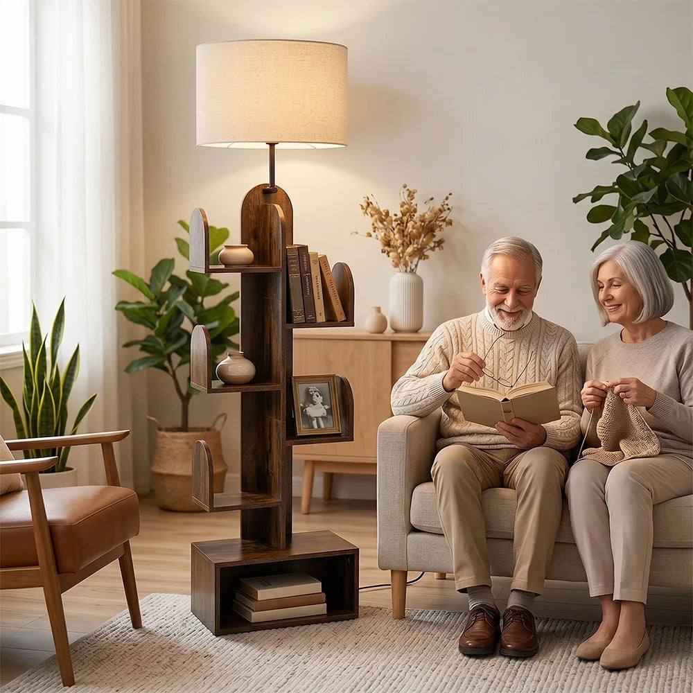 A tall, dark wood floor lamp with a beige drum shade stands beside a sofa in a cozy living room. An older man sits on the left reading a book, and an older woman sits on the right knitting, creating a warm, domestic scene.