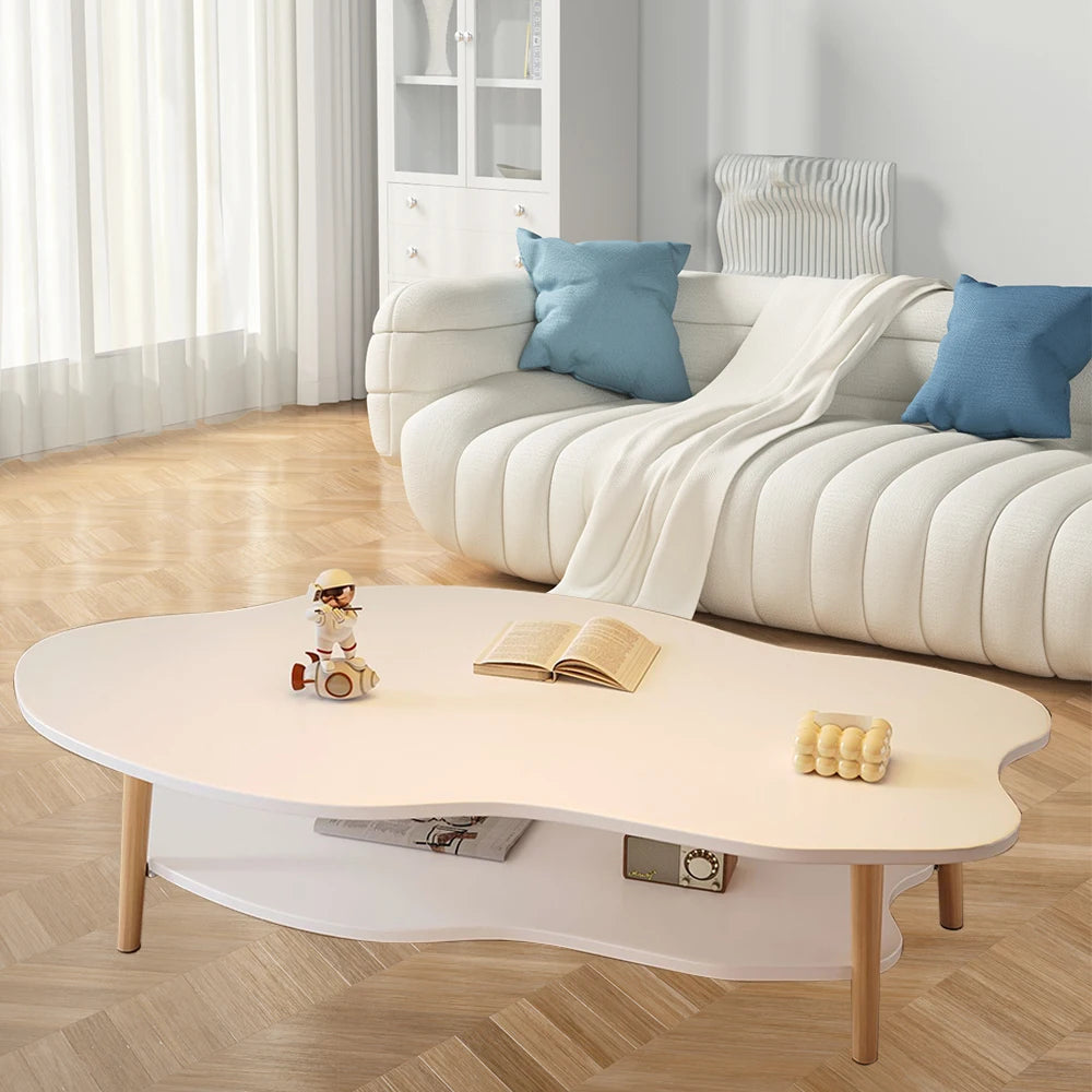 A modern organic-shaped white coffee table with light wood legs and a lower shelf, styled in a bright living room with a white curved sofa and blue pillows in the background.