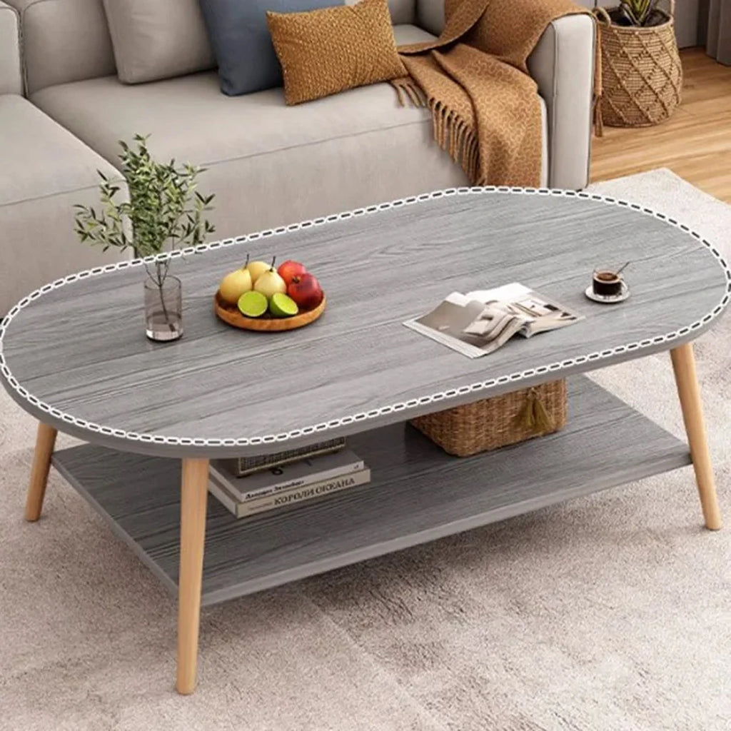 An oval gray coffee table with a white chain-link style border, four light wood legs, and a lower shelf, styled in a living room setting with a sofa, fruit, books, and a cup of coffee.