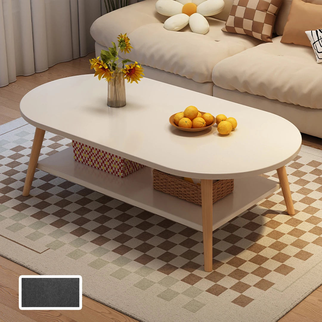 An oval-shaped white coffee table with light wood legs and a lower shelf, styled in a living room setting with a checkered rug, a sofa, and decorative items on top.
