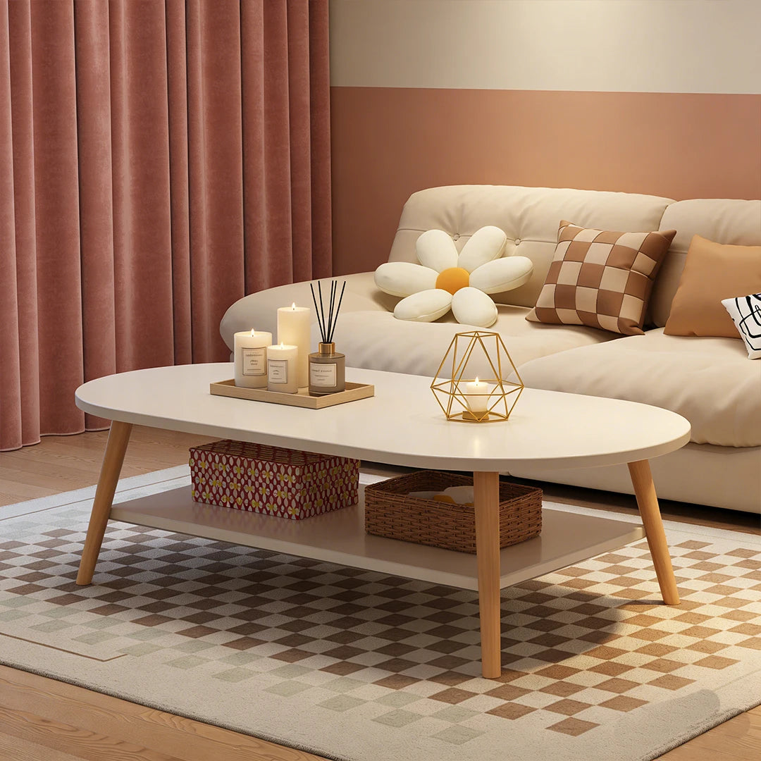 An oval coffee table with a white top and light wood legs in a styled living room. The table has a lower shelf with woven baskets and is decorated with candles and a geometric candle holder.