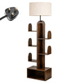 A modern dark brown wooden floor lamp with an arched trunk design, five tiered shelves, and a cylindrical off-white fabric drum shade. A black three-pin power plug is shown in the top-left corner against a solid white background.