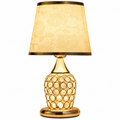 A table lamp with a warm gold-finished base featuring a circular crystal-like pattern, a matching gold-toned neck and base, and a tapered beige fabric shade with a subtle floral pattern and gold trim.
