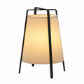 A modern table lamp with a black metal frame and a warm beige fabric drum shade, shown illuminated against a plain white background.