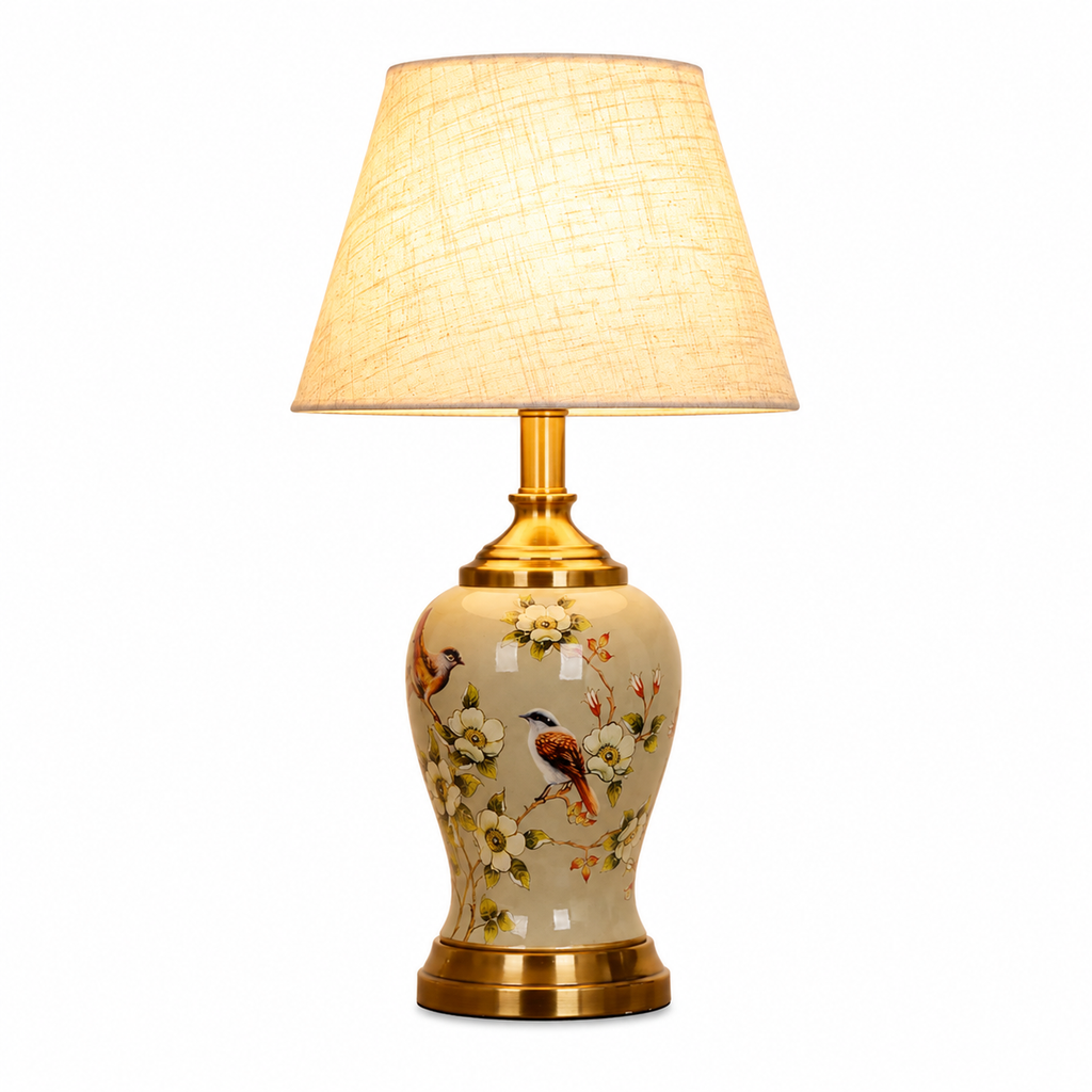 A table lamp with a warm beige fabric drum shade, a gold-finished neck and base, and a ceramic body decorated with birds and white blossoms, shown against a plain white background.