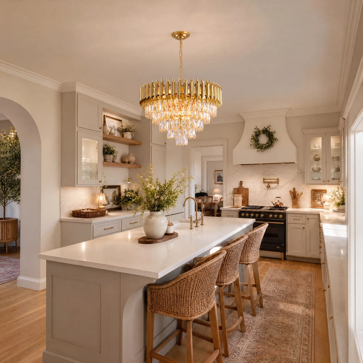 A luxury modern kitchen featuring a large gold chandelier with crystal droplets, a central island with a white countertop, three woven bar stools, and light wood flooring.