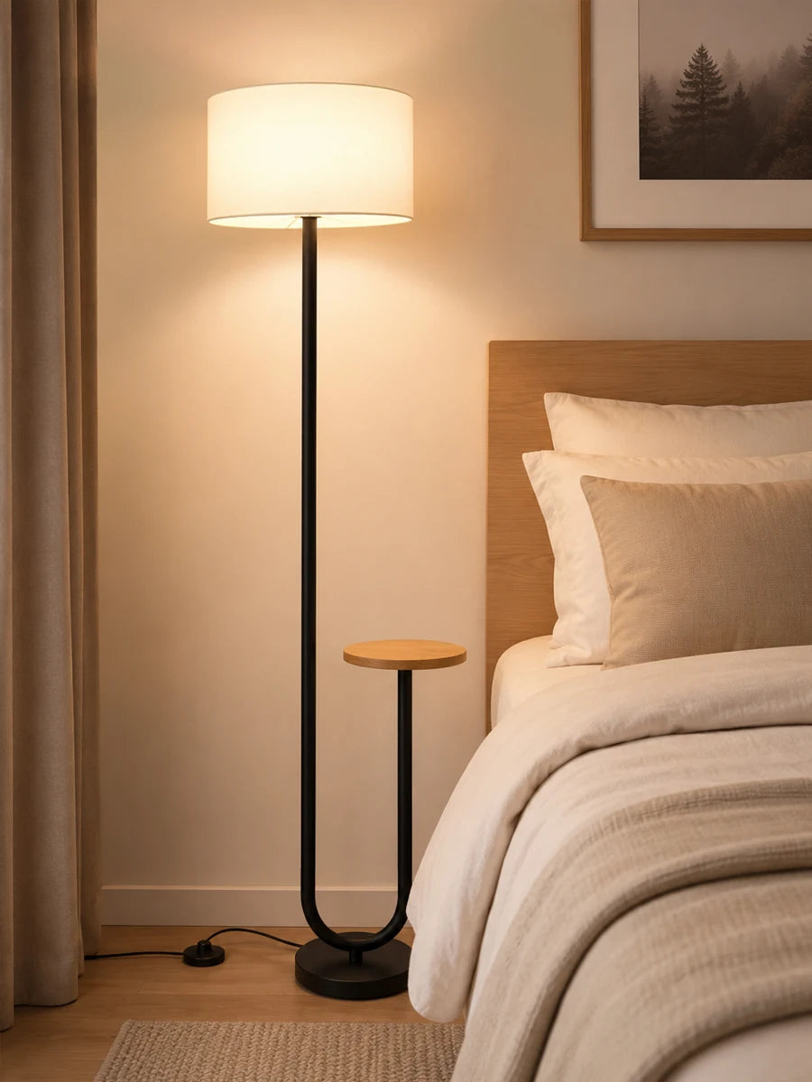 A modern floor lamp with a black metal stand and a warm white drum shade stands beside a neatly made bed in a softly lit bedroom.