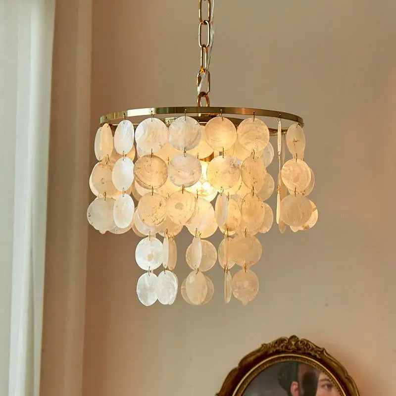 A hanging chandelier with a circular gold-toned frame and multiple dangling disc-shaped glass shades, illuminated in a warm light against an indoor wall.