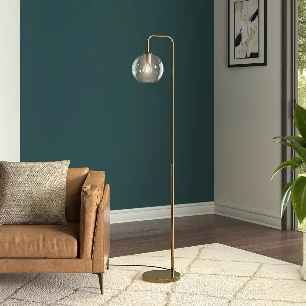 A modern floor lamp with a slim brass-colored metal stand and a clear glass globe shade, positioned in a living room next to a brown leather armchair against a dark teal wall.