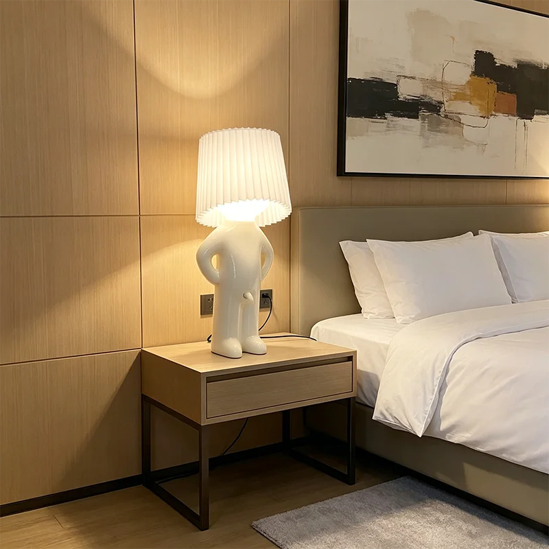 A modern white sculptural table lamp with a pleated white shade sits on a light wood nightstand beside a bed in a contemporary bedroom with wood-paneled walls.