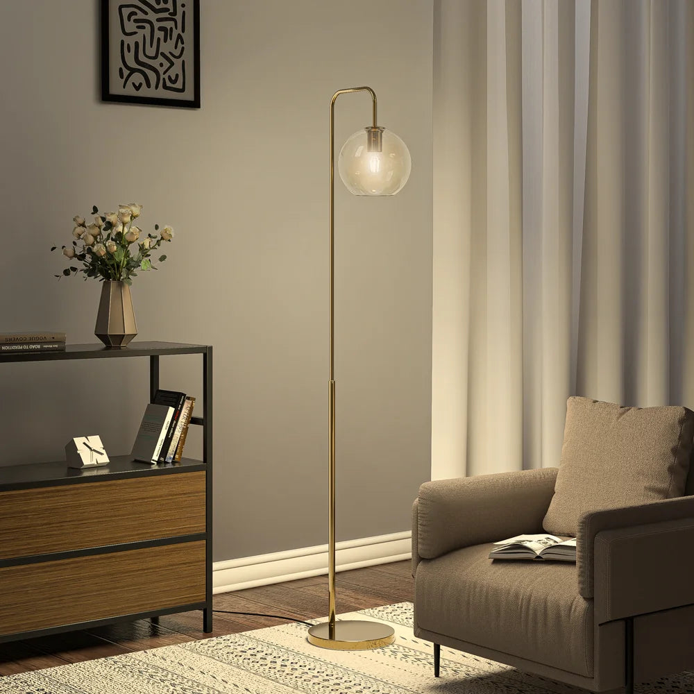 A modern floor lamp with a slim gold-finished metal stand and a round frosted glass globe shade, positioned in a styled living room next to a beige armchair and a wooden sideboard.