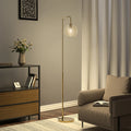 A modern floor lamp with a slim gold-finished metal stand and a round frosted glass globe shade, positioned in a styled living room next to a beige armchair and a wooden sideboard.