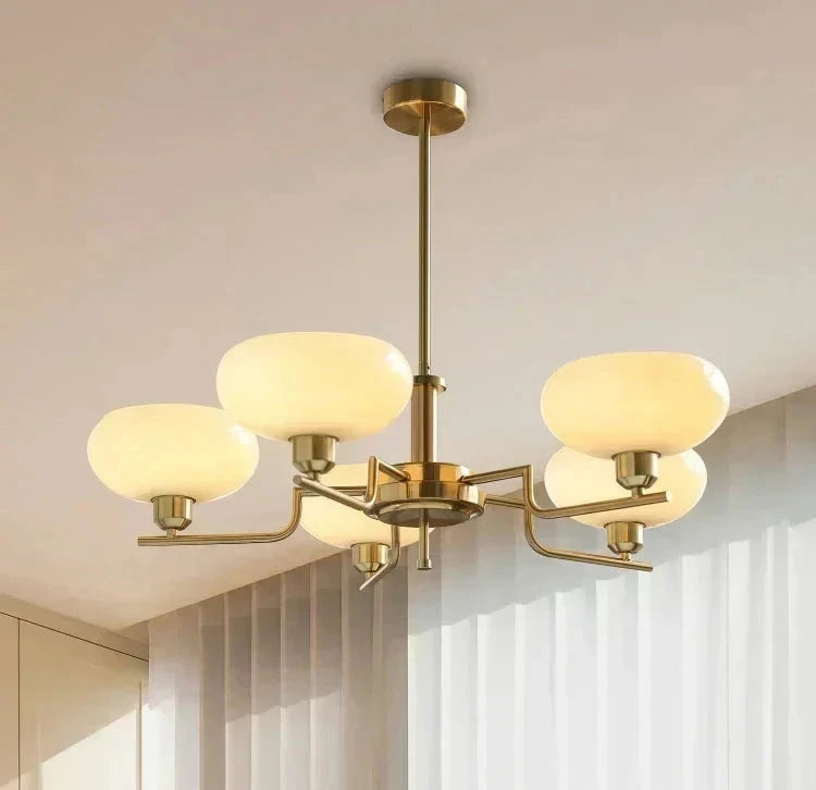 A modern five-light chandelier with a brushed brass/gold metal frame and oval frosted glass shades, hanging from a ceiling in a bright, contemporary room with white vertical blinds.