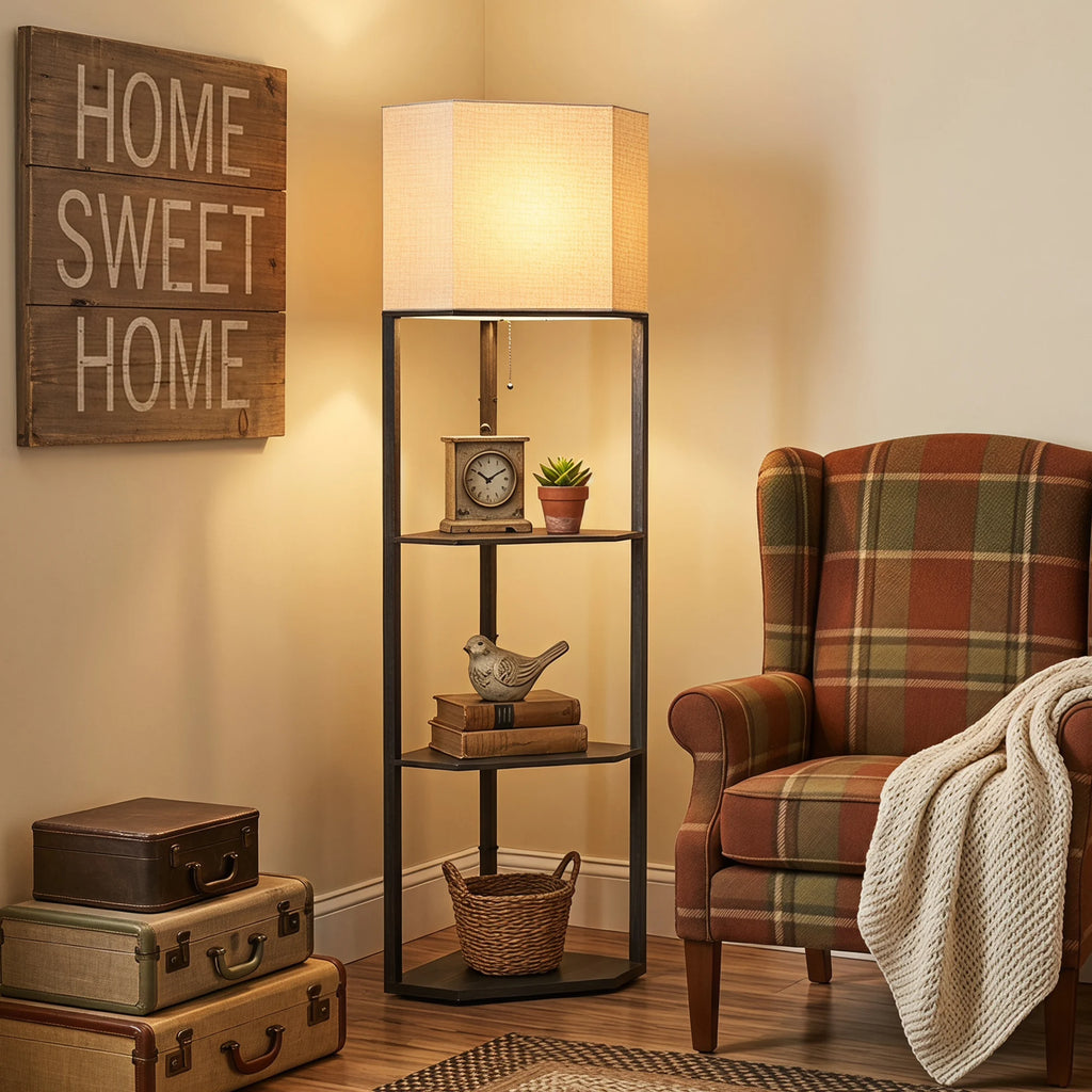 A tall black metal floor lamp with a warm beige fabric shade stands in a cozy room corner beside a plaid armchair, with decorative books, a small clock, a bird figurine, a basket, and stacked vintage suitcases against a cream wall with a wooden 'HOME SWEET HOME' sign.