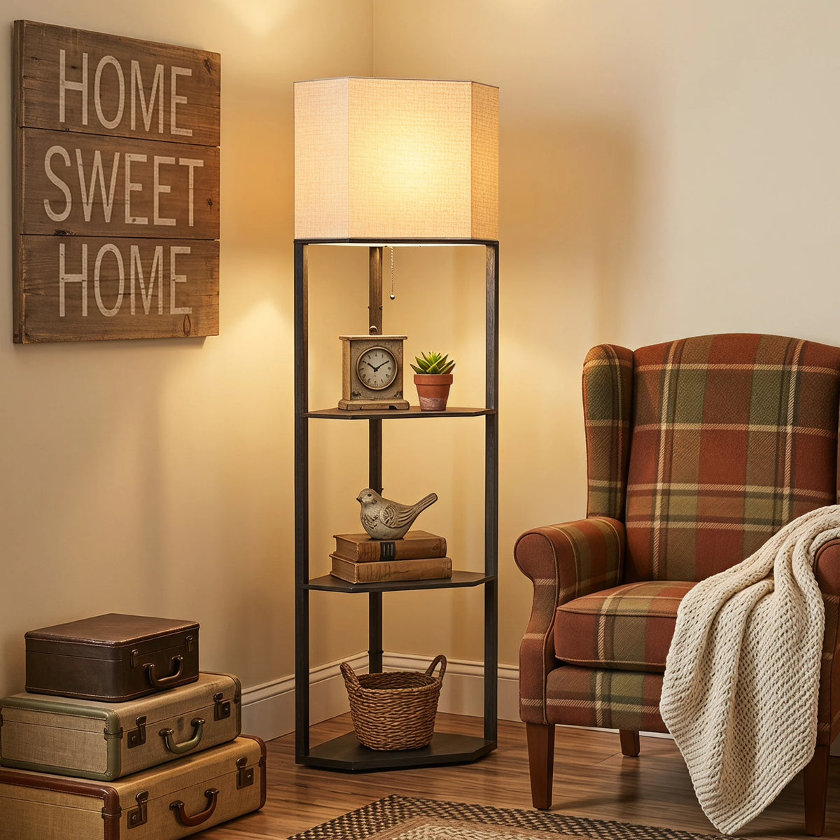 A tall black metal floor lamp with a warm beige fabric shade stands in a cozy room corner beside a plaid armchair, with decorative books, a small clock, a bird figurine, a basket, and stacked vintage suitcases against a cream wall with a wooden 'HOME SWEET HOME' sign.