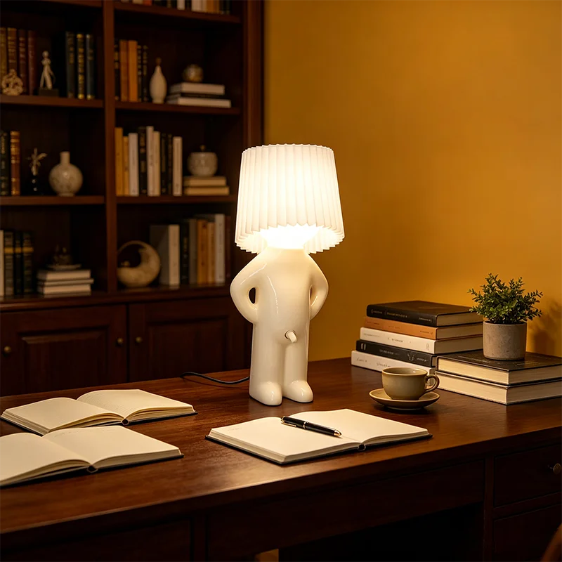A modern sculptural table lamp shaped like a white human figure with a pleated white lampshade head, placed on a wooden desk in a warmly lit home office setting.