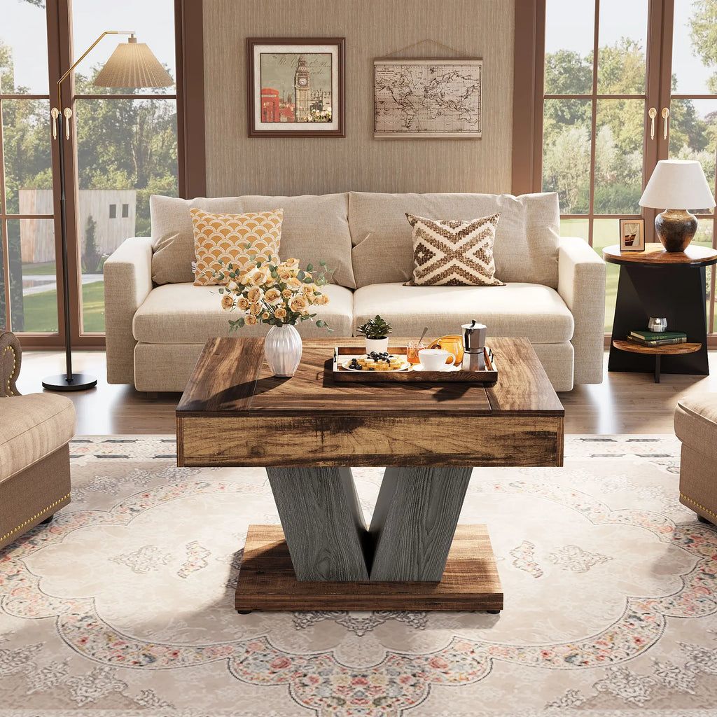 A rustic wooden coffee table with a V-shaped gray wood base is centered in a bright, modern living room. The table is styled with a matching wooden tray holding a teapot, cups, and a small vase of flowers, set on a large beige patterned rug.