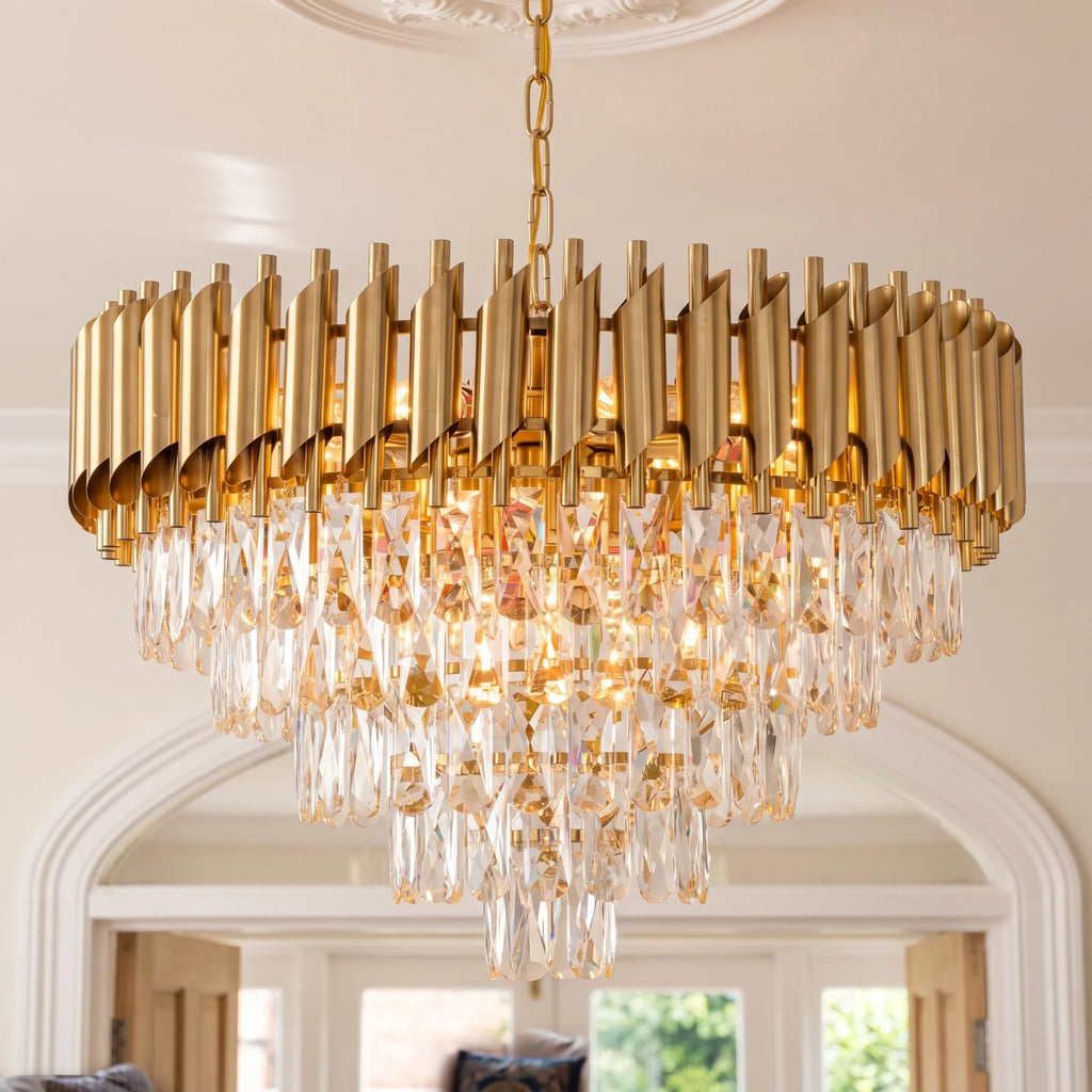 A modern gold chandelier with a circular frame made of vertical brushed metal bars and cascading clear crystal droplets, hanging in an elegant interior room with arched doorways.