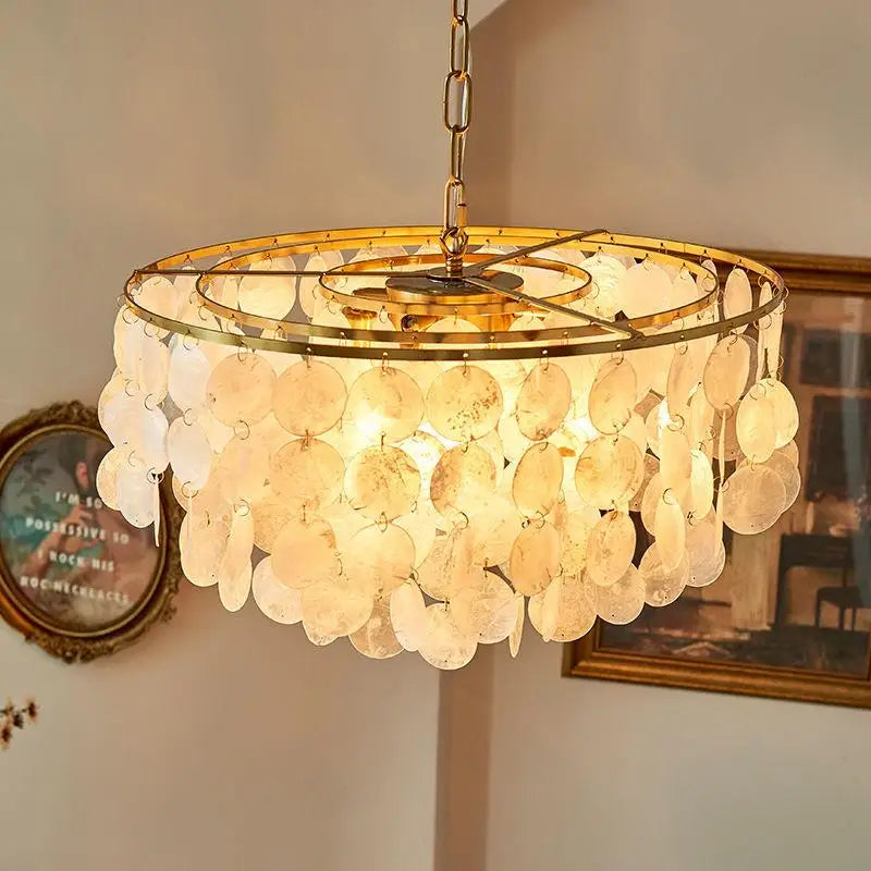 A round pendant chandelier with a gold-toned metal frame and numerous translucent, disc-like shell or glass drops, illuminated from within in a warm light, hanging in an indoor room setting.