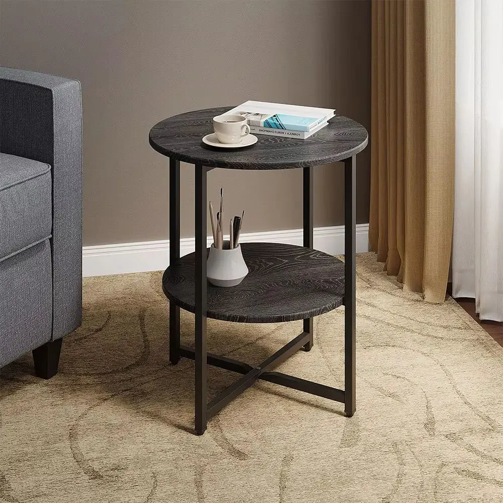 A round two-tier end table with a dark gray wood grain finish and black metal legs, styled in a living room next to a gray armchair on a beige patterned rug.
