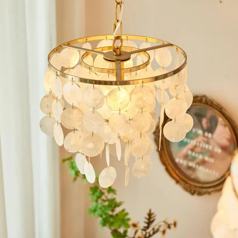 A hanging chandelier with a gold-toned circular frame and many translucent shell-like discs that glow warm white when lit, shown in a softly styled interior setting.