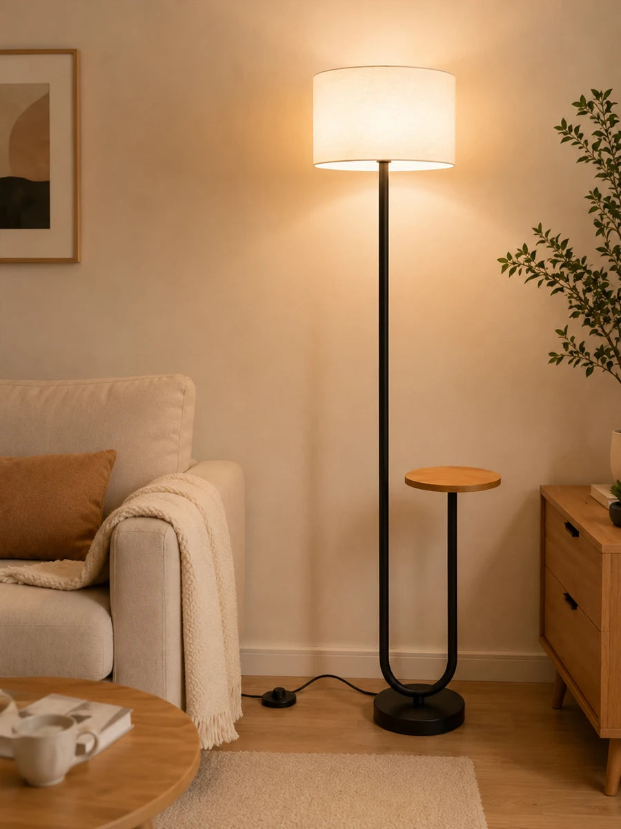 A modern floor lamp with a tall black metal stand and a cylindrical white shade, glowing with warm light in a cozy living room next to a beige sofa and wooden side table.