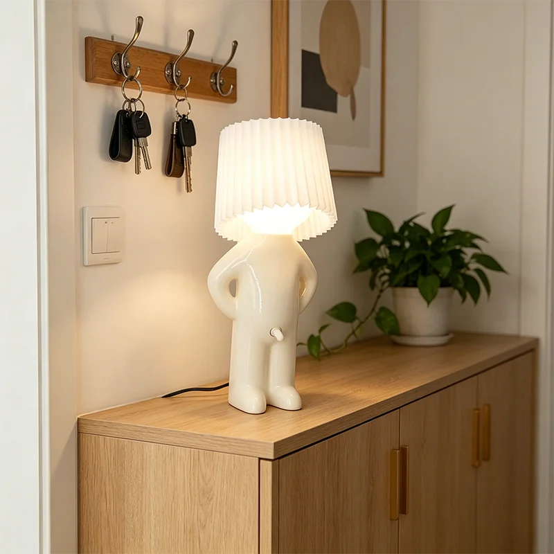 A white sculptural table lamp shaped like a person standing with hands on hips, with a pleated lampshade as the head, placed on a light wood cabinet in a home entryway.