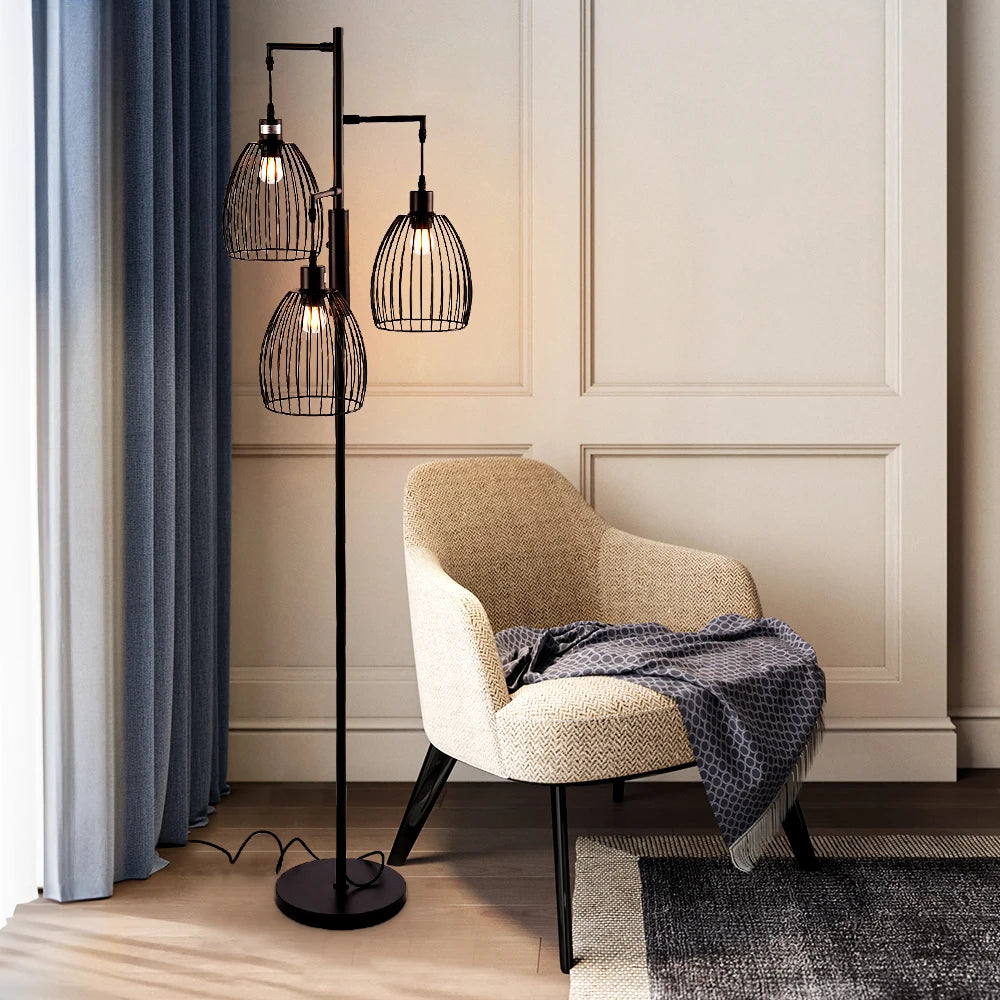 Modern floor lamp with three glass shades next to a chair in a room with neutral walls and curtains.