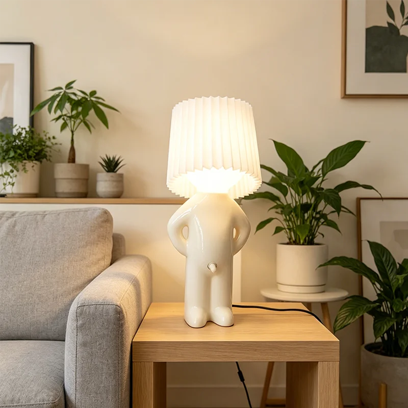 A white sculptural table lamp shaped like a person with hands on hips, featuring a pleated drum shade, displayed on a wooden side table in a cozy living room setting.