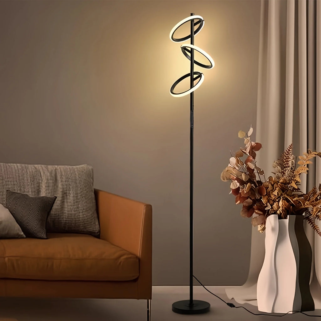 A modern spiral LED floor lamp with a black metal frame and glowing white light rings, shown in a styled living room next to an orange-brown sofa and a white vase with dried flowers.