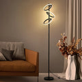 A modern spiral LED floor lamp with a black metal frame and glowing white light rings, shown in a styled living room next to an orange-brown sofa and a white vase with dried flowers.