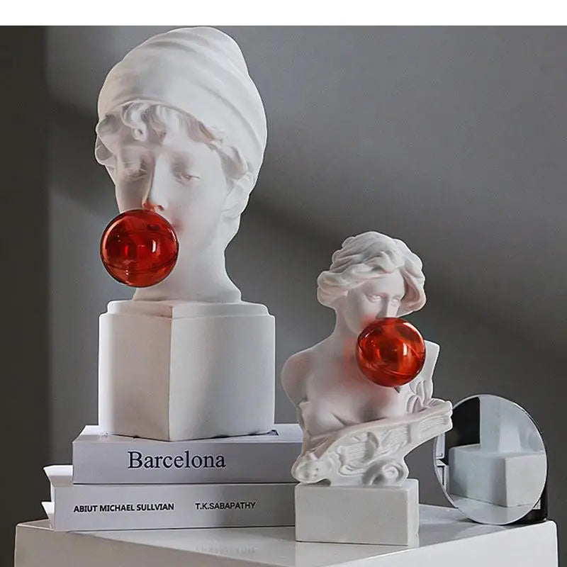 Two white classical-style bust sculptures light with glossy red orb accents, displayed on stacked books with a small round mirror on a gray background.