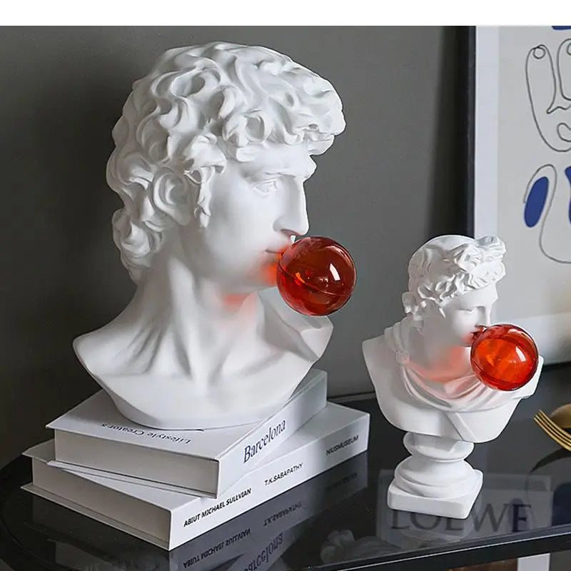 Two white classical bust sculpture lights on a glossy black table, each with a translucent red orb protruding from its mouth, styled with books against a dark gray wall.