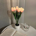 Potted plant like table lamp with glowing pink flowers on a table next to an open book and a glass.