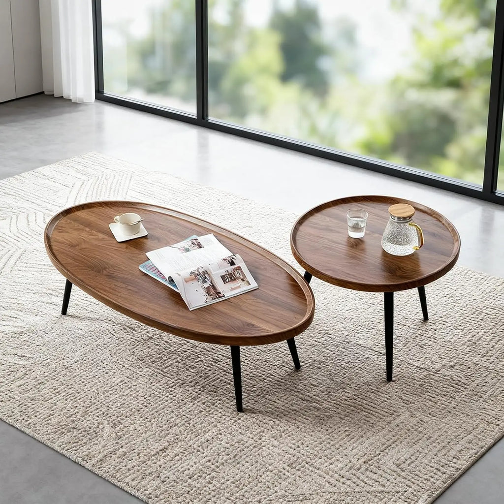 Two round wooden coffee tables with black metal legs, styled on a textured beige rug in a bright room with large windows. One table is oval-shaped and holds an open magazine and a small cup, while the other round table holds a glass cup and a glass pitcher.