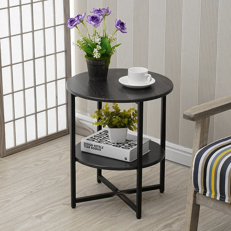 A round black metal side table with two shelves, styled with a potted purple flower on top, a white cup and saucer, and a decorative book on the shelf below, set in a bright room next to a window and a striped chair.