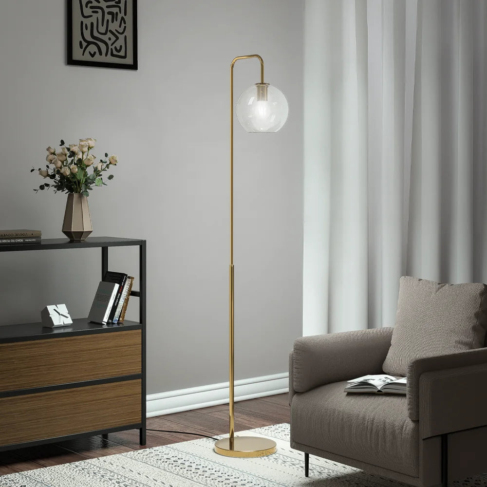 A modern floor lamp with a slim gold metal stand and a frosted white globe shade stands in a styled living room next to a gray armchair and a black-and-wood sideboard.