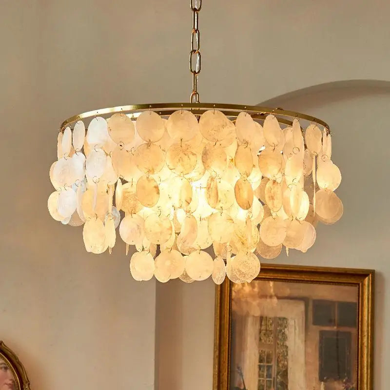 A round chandelier with a gold-tone metal frame and cascading shell-like discs that glow warm white when lit, hanging in an indoor room with a beige wall and framed artwork in the background.