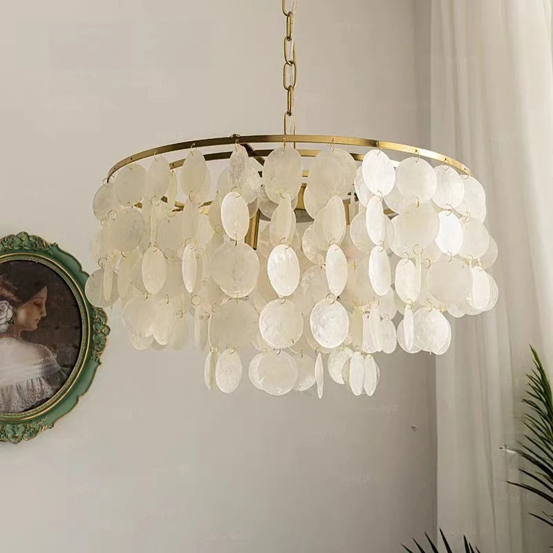 A modern chandelier with a gold-toned circular frame and multiple layers of textured, disc-like white shell pendants, hanging in a styled interior setting.