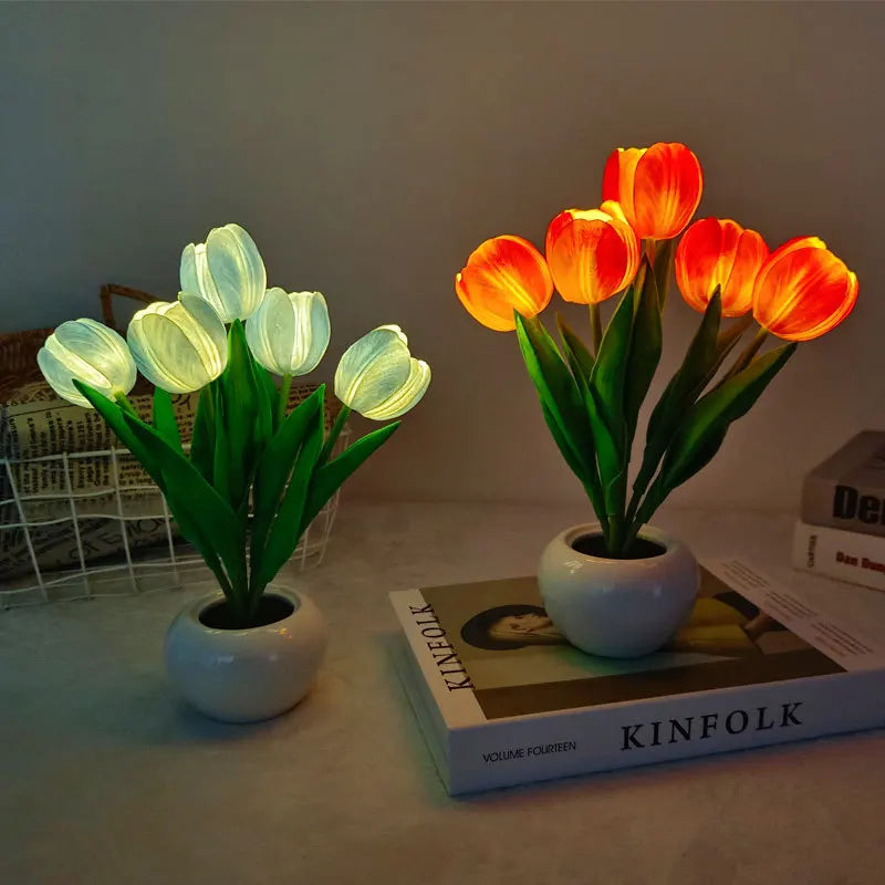 Two potted tulip lights, one glowing green and the other glowing orange table lamp, on a surface with a magazine underneath.